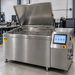 A large stainless steel industrial hot water bath or deep fryer with its lid open, featuring two wire baskets inside and a digital control panel on the right side, set in a clean workshop environment.